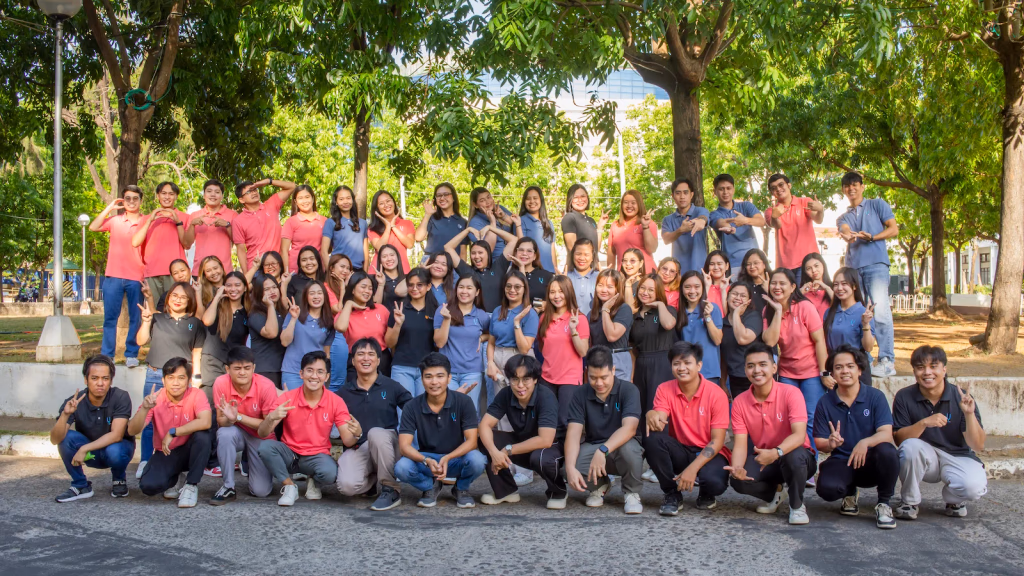 A large group of people in matching pink, blue, and black polo shirts posing for a group photo outdoors in front of lush green trees.