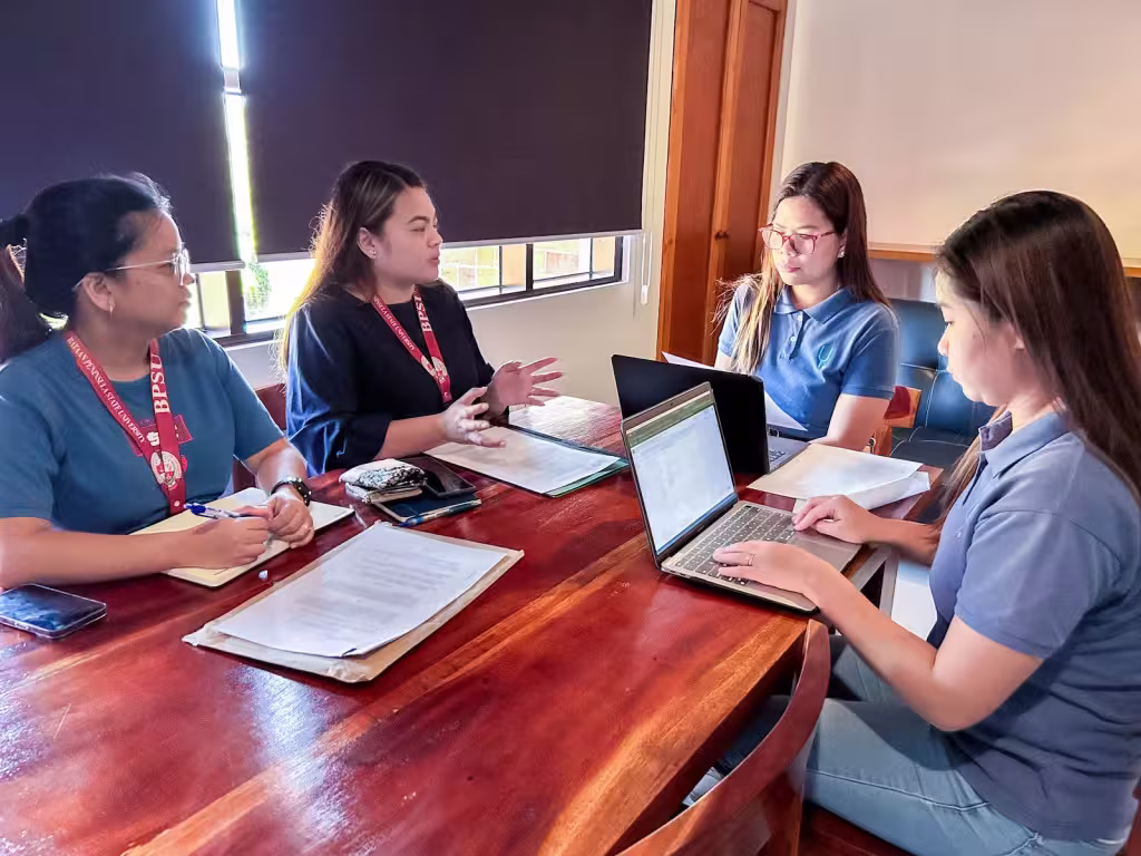 Four women sitting around a wooden table in an office setting, engaged in a meeting with laptops and documents.