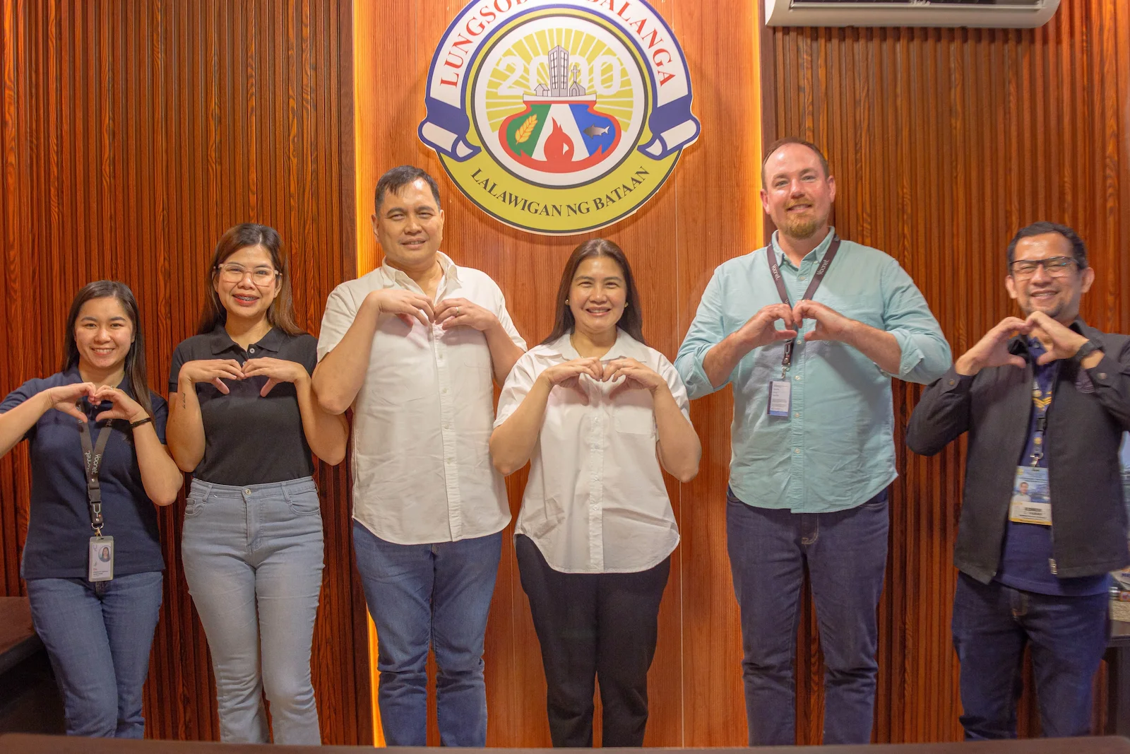 Six people standing in a row against a wood-paneled wall, all making heart shapes with their hands