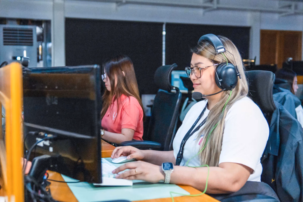 A woman wearing a headset and glasses works at a computer in an office setting, with a colleague visible in the background.