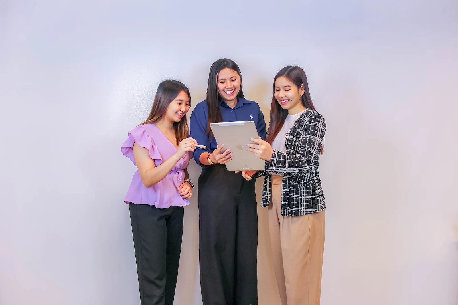 Three professional women smiling and collaborating while looking at a tablet together against a plain light gray background.